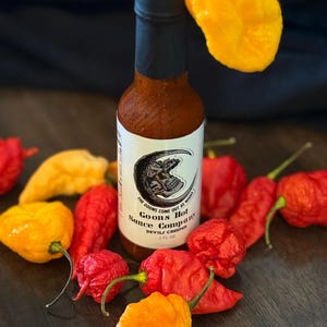 May include: A bottle of Goons Hot Sauce Company Devil's Creeper hot sauce with a black cap and label featuring a skull. The bottle is surrounded by red, orange, and yellow peppers on a dark wooden surface.