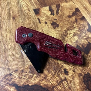 May include: A red utility knife with the brand name "Milwaukee" printed on the side. The knife has a black blade and a textured handle. The knife is designed for cutting and other utility tasks.