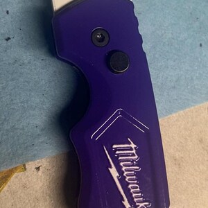 May include: A utility knife with a white blade and a purple handle. The handle has the Milwaukee logo and the word "FASTBACK" printed on it. The knife is designed for cutting and general utility tasks.