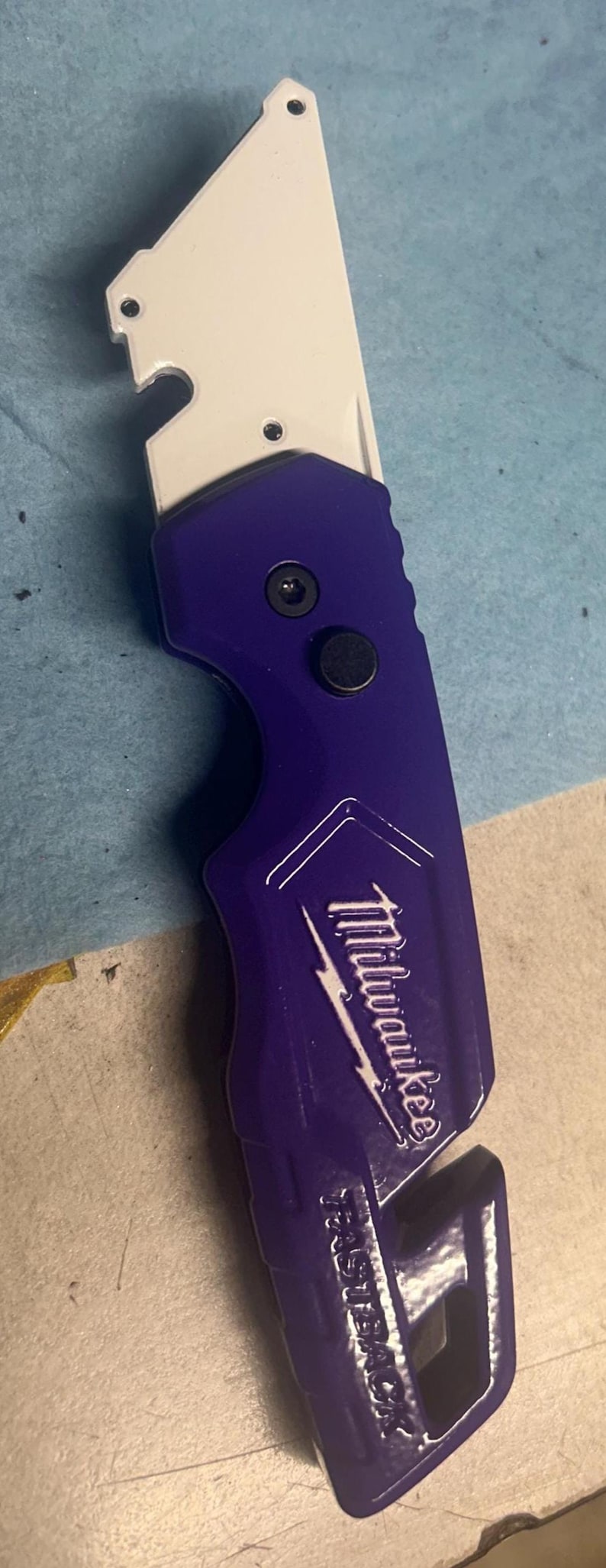 May include: A utility knife with a white blade and a purple handle. The handle has the Milwaukee logo and the word "FASTBACK" printed on it. The knife is designed for cutting and general utility tasks.