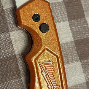 May include: Orange utility knife with a silver blade. The knife has the "Milwaukee" logo and "FASTBACK" text. The knife has two black buttons and a built-in bottle opener.