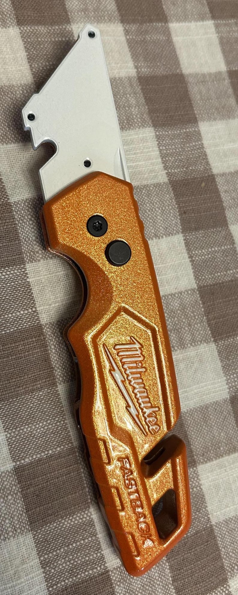 May include: Orange utility knife with a silver blade. The knife has the "Milwaukee" logo and "FASTBACK" text. The knife has two black buttons and a built-in bottle opener.