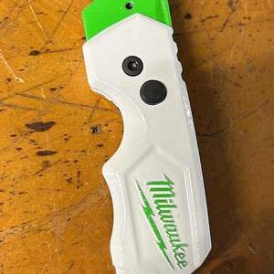 May include: A white and green utility knife with the brand name "Milwaukee" printed in green. The knife has a green blade and a white handle with a black button. The text "FASTBACK" is printed on the handle.