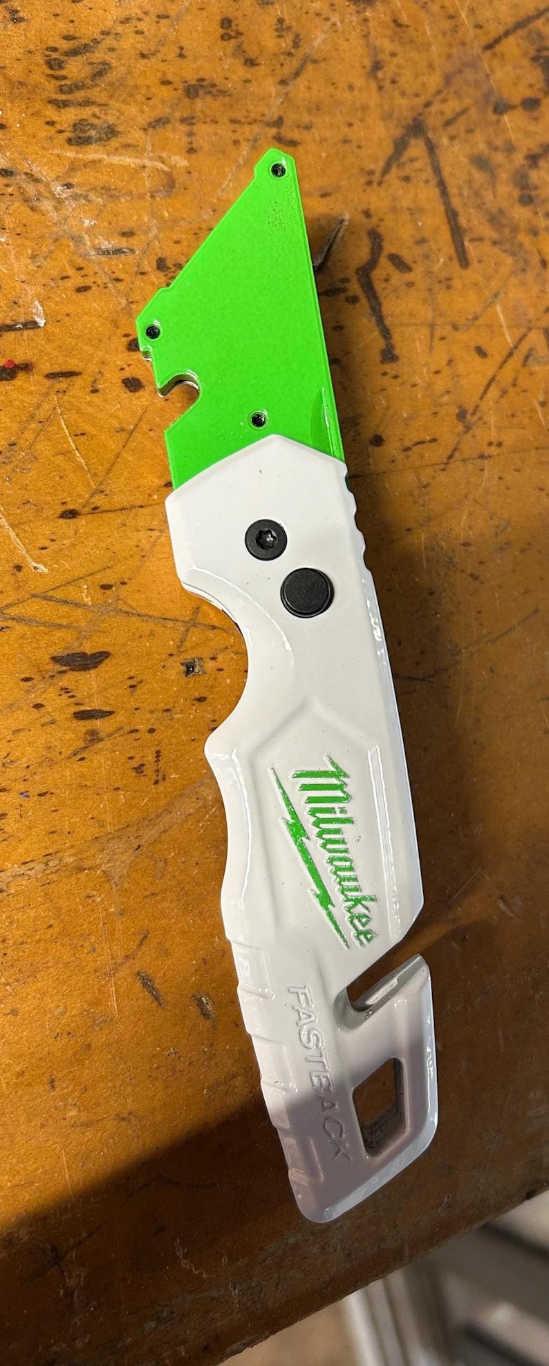 May include: A white and green utility knife with the brand name "Milwaukee" printed in green. The knife has a green blade and a white handle with a black button. The text "FASTBACK" is printed on the handle.