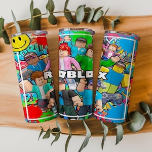 May include: Three cylindrical tumblers with metal lids, each featuring a vibrant Roblox-themed graphic print. The designs include various blocky character figures, game controllers, a yellow smiley face, and the word Roblox in bold white letters.