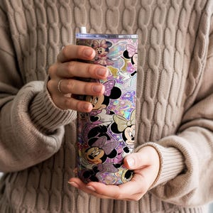 Op de afbeelding: Een cilindervormige beker met een herhalend patroon van Minnie Mouse-figuren in verschillende houdingen, gekleed in roze en paarse outfits, tegen een glinsterende, holografische en glitterende achtergrond met een doorzichtig deksel en een wit rietje.