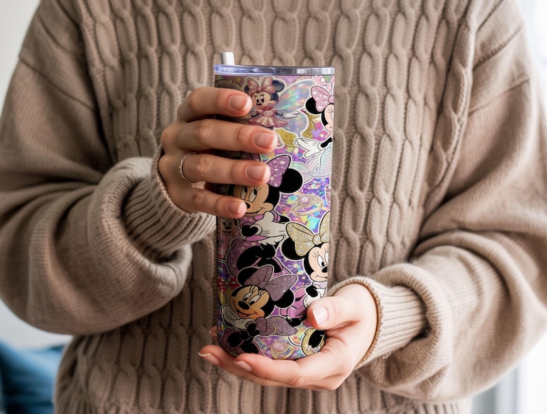 Op de afbeelding: Een cilindervormige beker met een herhalend patroon van Minnie Mouse-figuren in verschillende houdingen, gekleed in roze en paarse outfits, tegen een glinsterende, holografische en glitterende achtergrond met een doorzichtig deksel en een wit rietje.
