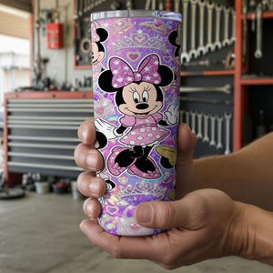 Op de afbeelding: Een cilindrische roestvrijstalen beker met een herhalend patroon van Minnie Mouse in een roze jurk met stippen en een strik, afgewisseld met glinsterende tiara's, hartjes en handtassen tegen een levendige paarse en roze holografische achtergrond.