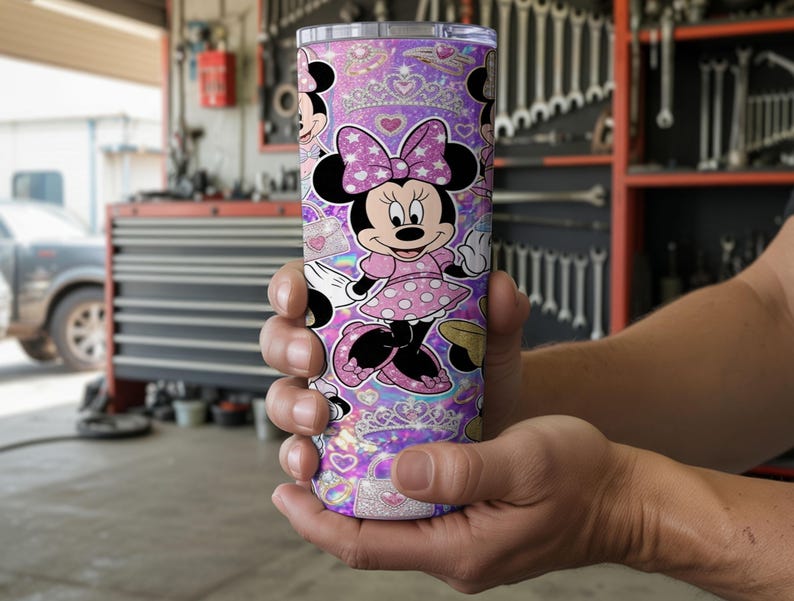 Op de afbeelding: Een cilindrische roestvrijstalen beker met een herhalend patroon van Minnie Mouse in een roze jurk met stippen en een strik, afgewisseld met glinsterende tiara's, hartjes en handtassen tegen een levendige paarse en roze holografische achtergrond.