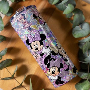 Op de afbeelding: Een cilindrische roestvrijstalen beker met een levendige, holografische wikkel met meerdere illustraties van Minnie Mouse in verschillende houdingen, gekleed in paarse en roze outfits, geaccentueerd door glinsterende glitter en fee-achtige vleugels.