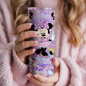 Op de afbeelding: Een cilindrische roestvrijstalen beker met een paarse glitterachtergrond en een herhalend patroon van Minnie Mouse, sprankelende tiara's, handtassen en hartvormen. Het ontwerp bevat een doorzichtig deksel en een gladde afwerking.