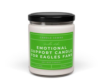 Può includere: Una candela di cera di soia bianca in un barattolo di vetro con coperchio nero. L'etichetta verde riporta la scritta "CANDLE JAWNS" e "EMOTIONAL SUPPORT CANDLE FOR EAGLES FANS". La candela è da 255g e dura 40 ore.