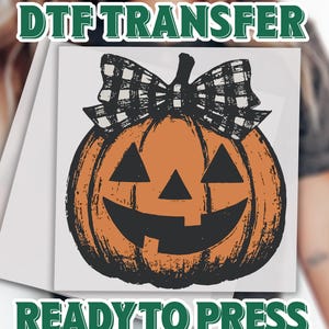 May include: A DTF transfer sheet featuring a graphic of an orange jack-o'-lantern pumpkin with a black and white buffalo plaid bow on top. The text DTF TRANSFER is printed in green above the graphic, and READY TO PRESS is printed in green below.
