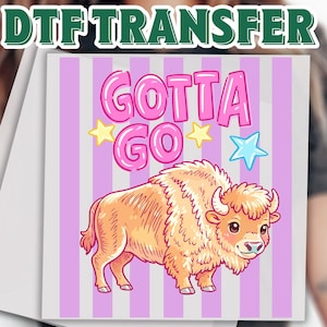 May include: A DTF transfer with the text "GOTTA GO" in pink bubble letters, a cartoon bison, and the text "READY TO PRESS". The bison is light brown with pink accents, set against a pink and white striped background.