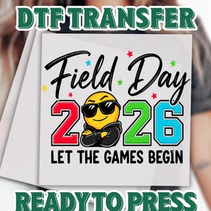 May include: A white DTF transfer with the text "Field Day 2026" and "Let the Games Begin". The design features a smiling emoji wearing sunglasses and a hoodie. The numbers 2 and 6 are red and blue, respectively.