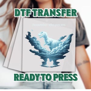May include: A DTF transfer featuring a blue Tyrannosaurus Rex illustration surrounded by stylized clouds. The text "Remember who you are" is above the dinosaur. The words "DTF TRANSFER" and "READY TO PRESS" are also displayed.