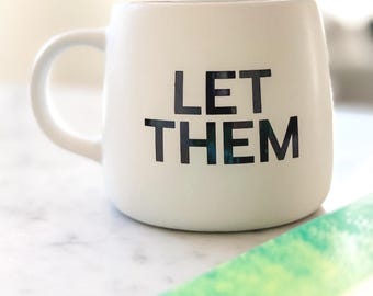 Könnte beinhalten: Eine weiße Keramik-Tasse mit gebogenem Henkel. Die Tasse trägt den schwarzen Text "LET THEM" in einer fetten, serifenlosen Schrift. Die Tasse steht auf einer weißen Oberfläche, mit einem Hauch von grünem Objekt im Hintergrund.