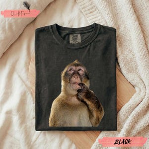 May include: Black t-shirt featuring a thoughtful monkey graphic. The monkey is depicted with its finger on its chin, suggesting contemplation. The shirt is labeled "BLACK" and has a "COMFORT COLORS" tag.