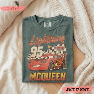 May include: Blue spruce t-shirt featuring a graphic of Lightning McQueen from the Cars movie. The design includes the number 95, a checkered flag, and the name "McQueen" in a vintage style. The shirt is folded on a wooden surface.