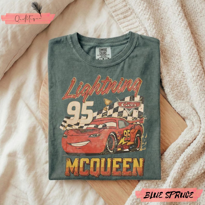 May include: Blue spruce t-shirt featuring a graphic of Lightning McQueen from the Cars movie. The design includes the number 95, a checkered flag, and the name "McQueen" in a vintage style. The shirt is folded on a wooden surface.