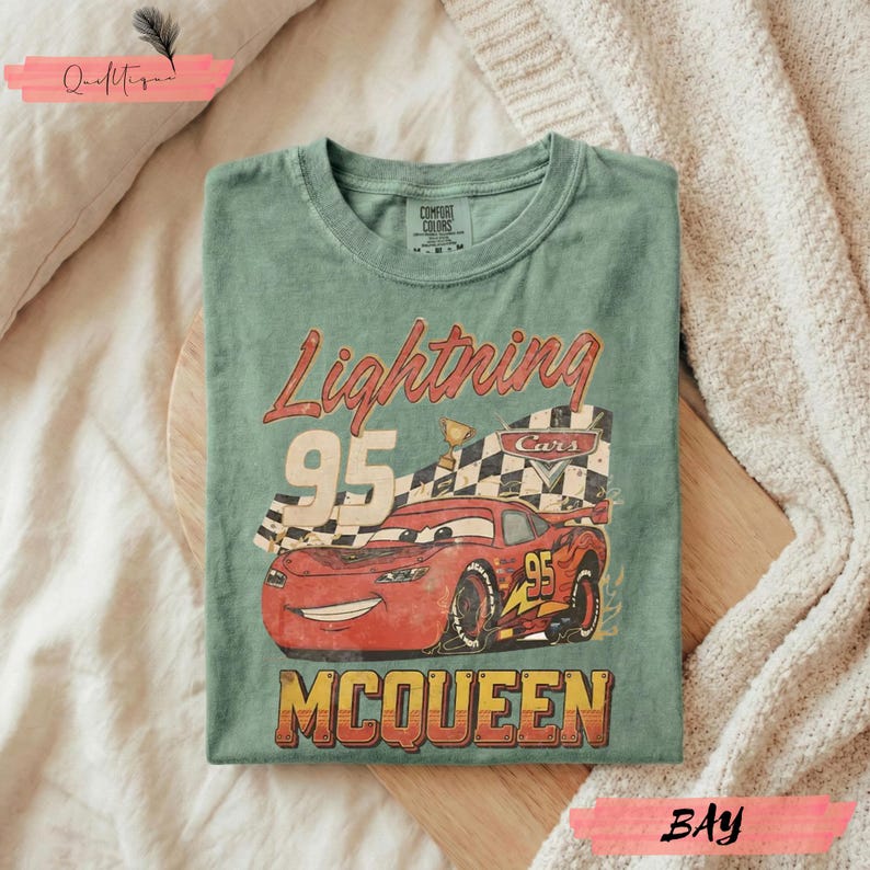 May include: Green t-shirt featuring a graphic of Lightning McQueen from the Cars movie. The design includes the text "Lightning 95 McQueen" with a checkered flag and trophy. The shirt is folded on a wooden surface.