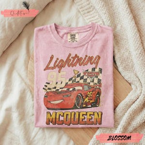 May include: Pink t-shirt featuring a graphic of Lightning McQueen from the Cars movie. The design includes the number 95, a checkered flag, and the name McQueen. The shirt is made of a soft material.
