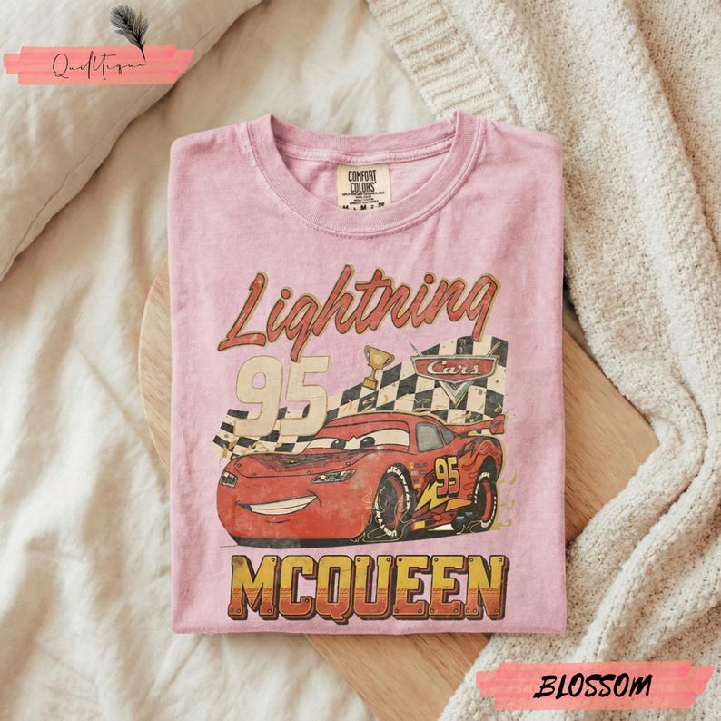 May include: Pink t-shirt featuring a graphic of Lightning McQueen from the Cars movie. The design includes the number 95, a checkered flag, and the name McQueen. The shirt is made of a soft material.