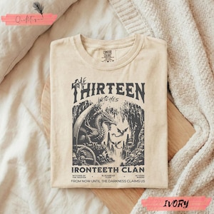 May include: Ivory t-shirt with a detailed graphic of a dragon, castle, and the text "The Thirteen Witches Ironteeth Clan." The design includes the phrases "Witches of the Wastes," "Blackbear Clan," and "Wyvern Riders."