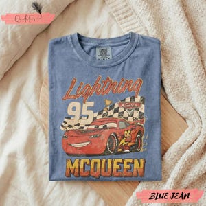 May include: Blue jean-colored t-shirt featuring a graphic of Lightning McQueen from the Cars movie. The design includes the text "Lightning 95 McQueen" with a checkered flag and a red race car. The shirt is folded on a wooden surface.
