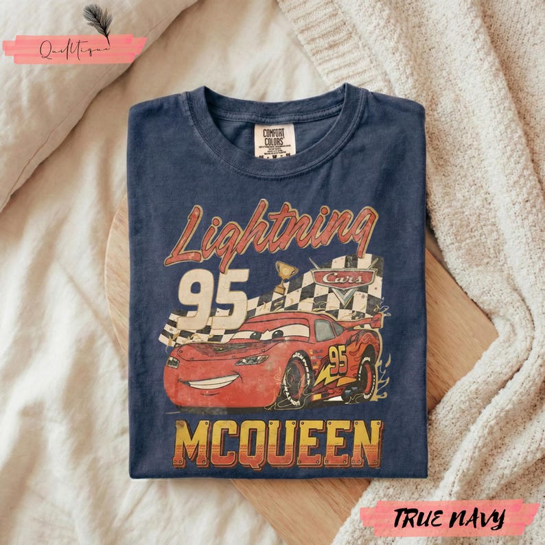 May include: Navy blue t-shirt featuring a graphic of Lightning McQueen from the Cars movie. The design includes the text "Lightning 95 McQueen" in a vintage style. The shirt is folded on a wooden surface.
