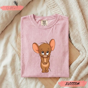 Puede incluir: Camiseta rosa claro con un ratón de dibujos animados de grandes orejas y una expresión dulce. La camiseta está doblada y tiene una etiqueta en el cuello. La palabra "BLOSSOM" está impresa en una pancarta rosa.