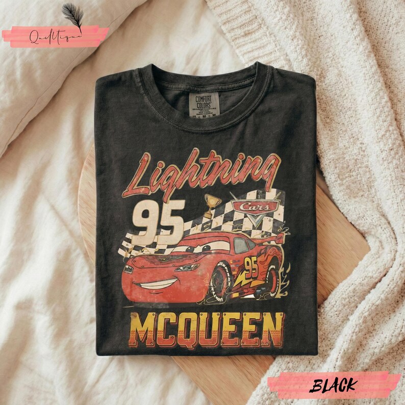 May include: Black t-shirt featuring a graphic of Lightning McQueen from the Cars movie. The design includes the text "Lightning 95 McQueen" in red and yellow, along with a checkered flag and a trophy. The shirt is folded.