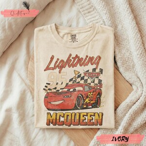 May include: Ivory-colored t-shirt featuring a graphic of Lightning McQueen from the Cars movie. The design includes the text "Lightning 95 McQueen" with a checkered flag and trophy. The shirt is folded on a wooden surface.