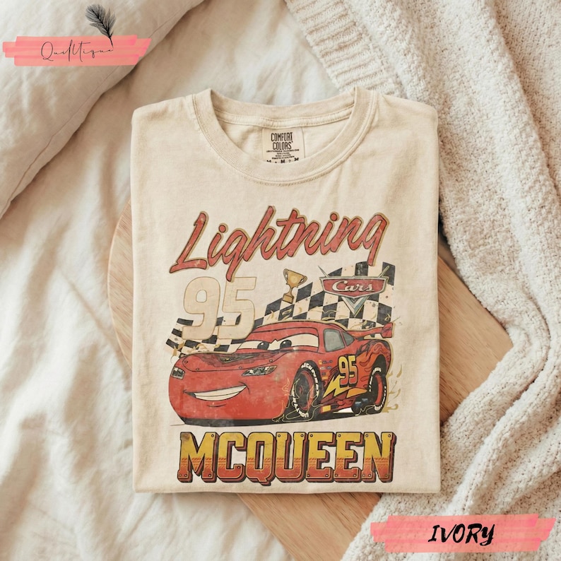 May include: Ivory-colored t-shirt featuring a graphic of Lightning McQueen from the Cars movie. The design includes the text "Lightning 95 McQueen" with a checkered flag and trophy. The shirt is folded on a wooden surface.