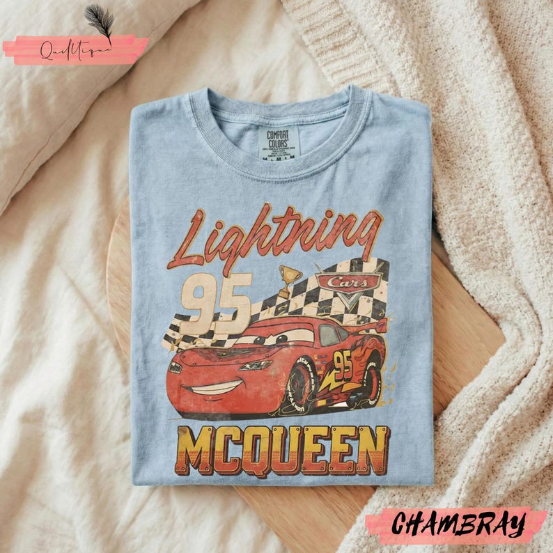 May include: Light blue t-shirt featuring a graphic of Lightning McQueen from the Cars movie. The design includes the text "Lightning 95 McQueen" in a vintage style. The shirt is folded on a wooden surface.