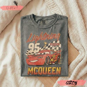 May include: A grey t-shirt featuring a graphic of Lightning McQueen from the Cars movie. The design includes the text "Lightning 95 McQueen" and a checkered flag. The shirt is made of a soft material.