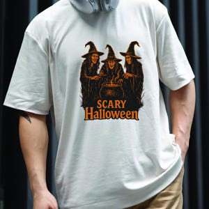 May include: Off-white t-shirt featuring a Halloween graphic. The design shows three witches around a cauldron with the words "Scary Halloween" in orange. The witches are wearing black robes and hats.