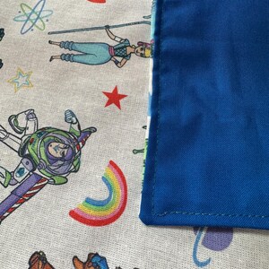 K&ouml;nnte beinhalten: Nahaufnahme eines Stoffes mit Toy Story-Motiv. Der Stoff zeigt Zeichentrickfiguren, eine rote Rakete, einen Regenbogen und einen blauen Rand. Die Hauptfarben sind Wei&szlig;, Blau, Rot und Gr&uuml;n.