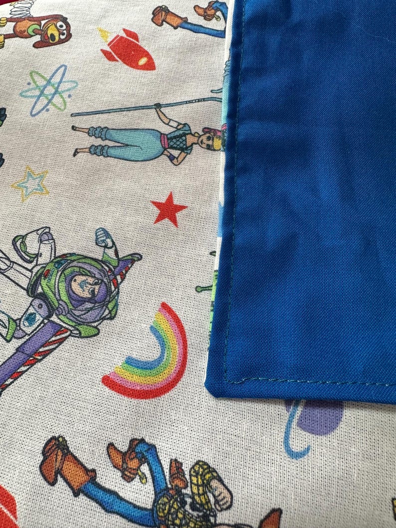 K&ouml;nnte beinhalten: Nahaufnahme eines Stoffes mit Toy Story-Motiv. Der Stoff zeigt Zeichentrickfiguren, eine rote Rakete, einen Regenbogen und einen blauen Rand. Die Hauptfarben sind Wei&szlig;, Blau, Rot und Gr&uuml;n.