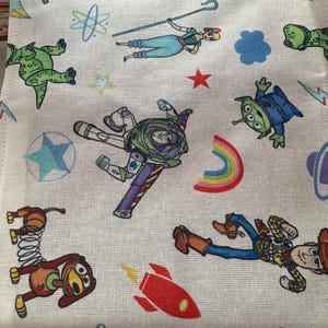 K&ouml;nnte beinhalten: Ein wei&szlig;er Stoff mit einem farbenfrohen Muster mit Figuren aus dem Film Toy Story. Das Design umfasst Buzz Lightyear, Woody, Bo Peep, Aliens, Raketen und andere verspielte Elemente. Der Stoff hat ein strukturiertes Aussehen.