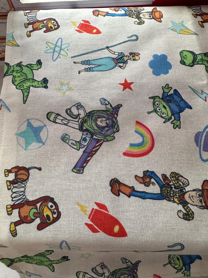K&ouml;nnte beinhalten: Ein wei&szlig;er Stoff mit einem farbenfrohen Muster mit Figuren aus dem Film Toy Story. Das Design umfasst Buzz Lightyear, Woody, Bo Peep, Aliens, Raketen und andere verspielte Elemente. Der Stoff hat ein strukturiertes Aussehen.