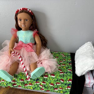 May include: A doll wearing a pink tutu dress, a red and white striped bow, and a red headband with white accents. The doll is sitting on a bed with a green and white patterned blanket and a white pillow. A candy cane is in the doll's hand.
