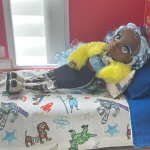 K&ouml;nnte beinhalten: Eine Puppe mit blauen Haaren und einer gelben Jacke liegt auf einem Bett mit einem blauen Kissen und einer gemusterten Decke mit Zeichentrickfiguren. Das Bett hat einen Holzrahmen. Der Hintergrund umfasst eine rote Wand und ein Fenster.