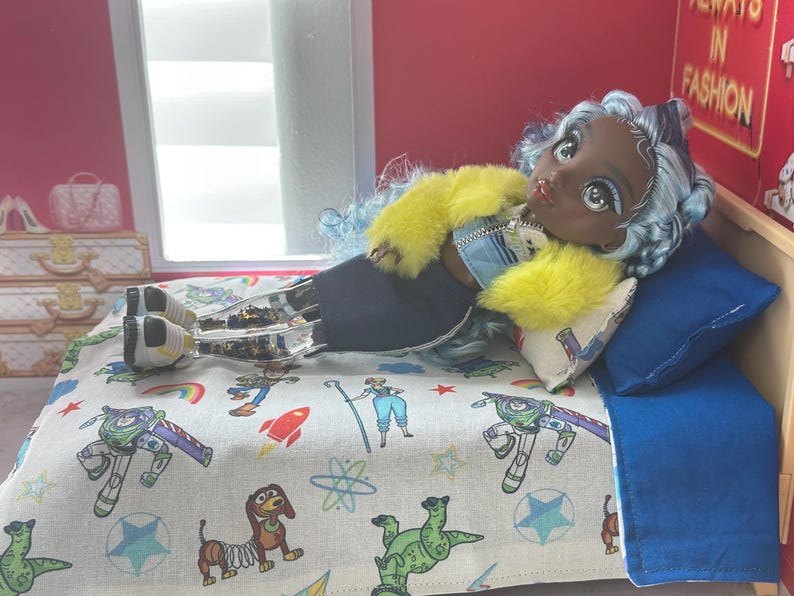 K&ouml;nnte beinhalten: Eine Puppe mit blauen Haaren und einer gelben Jacke liegt auf einem Bett mit einem blauen Kissen und einer gemusterten Decke mit Zeichentrickfiguren. Das Bett hat einen Holzrahmen. Der Hintergrund umfasst eine rote Wand und ein Fenster.