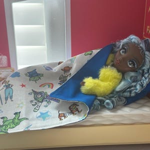 K&ouml;nnte beinhalten: Eine Puppe mit blauen Haaren und einem gelben, flauschigen Kleidungsst&uuml;ck liegt in einem Miniaturbett. Die Puppe ist mit einer Decke mit Zeichentrickfiguren bedeckt. Das Bett hat eine wei&szlig;e Matratze und ein blaues Kissen. Der Raum hat eine rosa Wand und ein Schild mit der Aufschrift "ALWAYS IN FASHION."