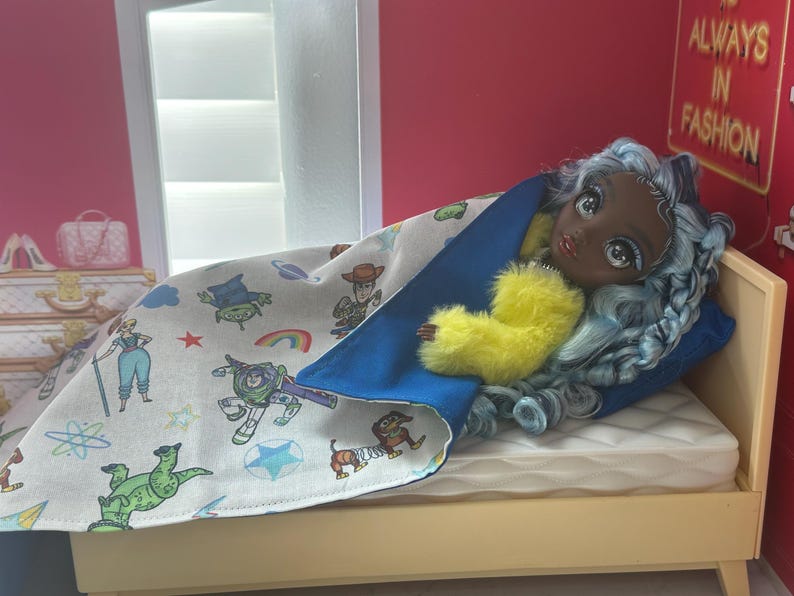 K&ouml;nnte beinhalten: Eine Puppe mit blauen Haaren und einem gelben, flauschigen Kleidungsst&uuml;ck liegt in einem Miniaturbett. Die Puppe ist mit einer Decke mit Zeichentrickfiguren bedeckt. Das Bett hat eine wei&szlig;e Matratze und ein blaues Kissen. Der Raum hat eine rosa Wand und ein Schild mit der Aufschrift "ALWAYS IN FASHION."