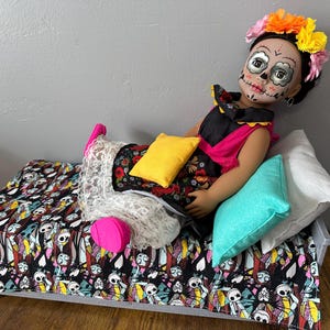 Peut inclure: Une poup&eacute;e avec un visage de cr&acirc;ne peint, portant une couronne de fleurs et une robe noire, rose et fleurie. La poup&eacute;e est allong&eacute;e sur un lit avec une couverture &agrave; motifs de cr&acirc;nes et de squelettes et plusieurs oreillers.