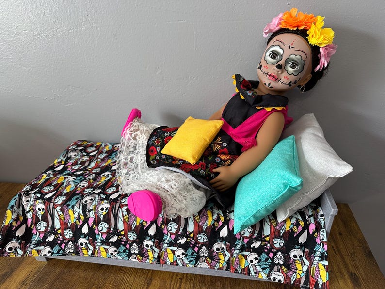 Peut inclure: Une poup&eacute;e avec un visage de cr&acirc;ne peint, portant une couronne de fleurs et une robe noire, rose et fleurie. La poup&eacute;e est allong&eacute;e sur un lit avec une couverture &agrave; motifs de cr&acirc;nes et de squelettes et plusieurs oreillers.