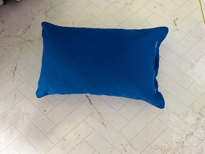 K&ouml;nnte beinhalten: Ein rechteckiges, einfarbig blaues Kissen. Das Kissen hat einen leuchtenden Blauton und scheint aus einem weichen Stoff zu bestehen. Das Kissen liegt auf einer hellen, gemusterten Oberfl&auml;che.