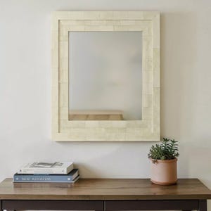 Könnte beinhalten: Ein rechteckiger Spiegel mit cremefarbenem Rahmen im Mosaikstil hängt an einer weißen Wand. Unter dem Spiegel zeigt ein Holztisch zwei Bücher und eine kleine Topfpflanze. Der Spiegel spiegelt einen Blick auf eine hellfarbene Bank wider.